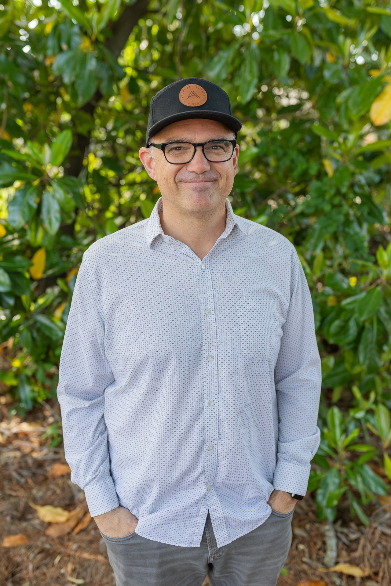 Person wearing glasses and a black cap stands in front of a leafy green background.