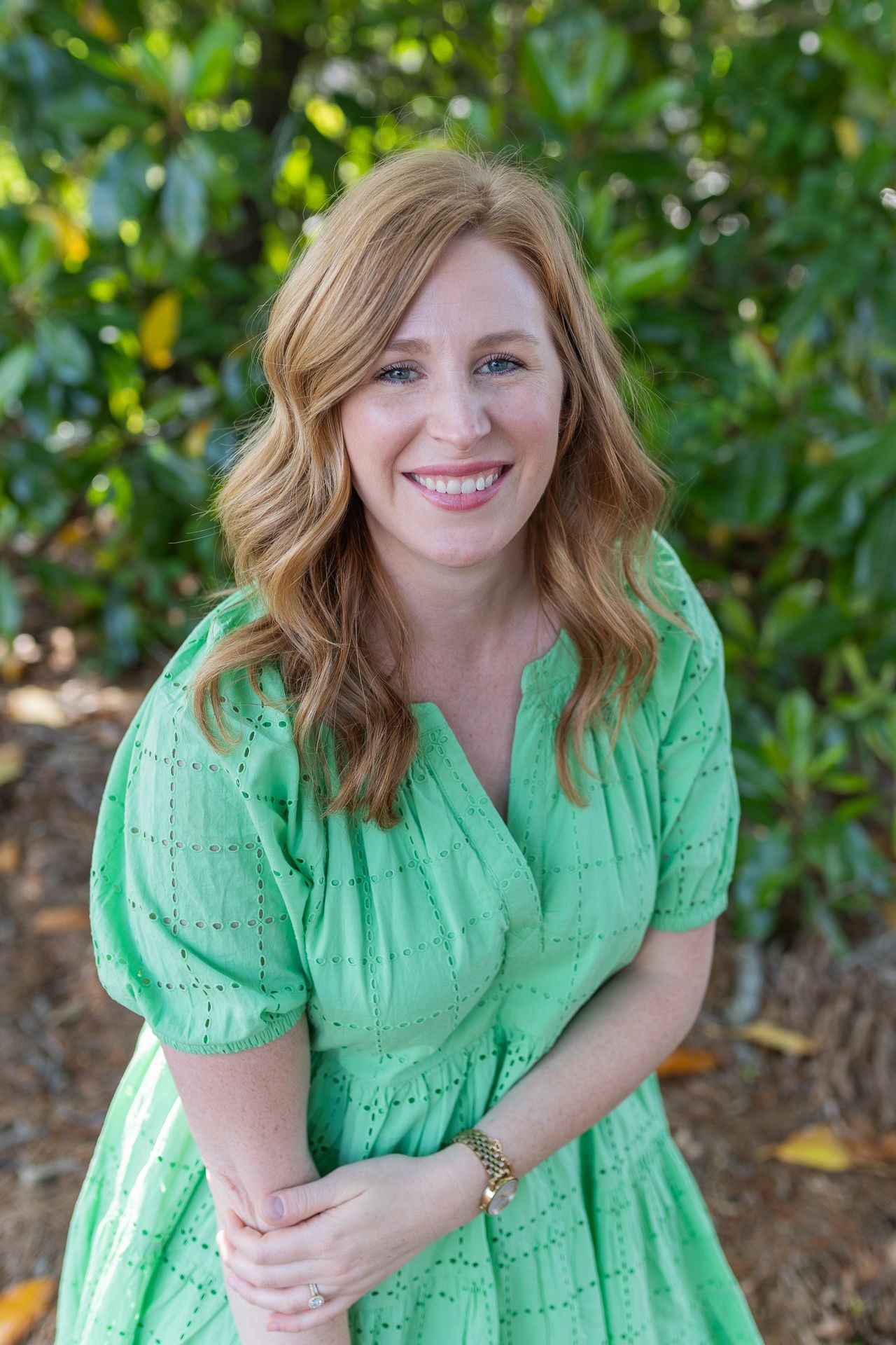 Person with wavy auburn hair wearing a green dress, standing against a leafy green background.