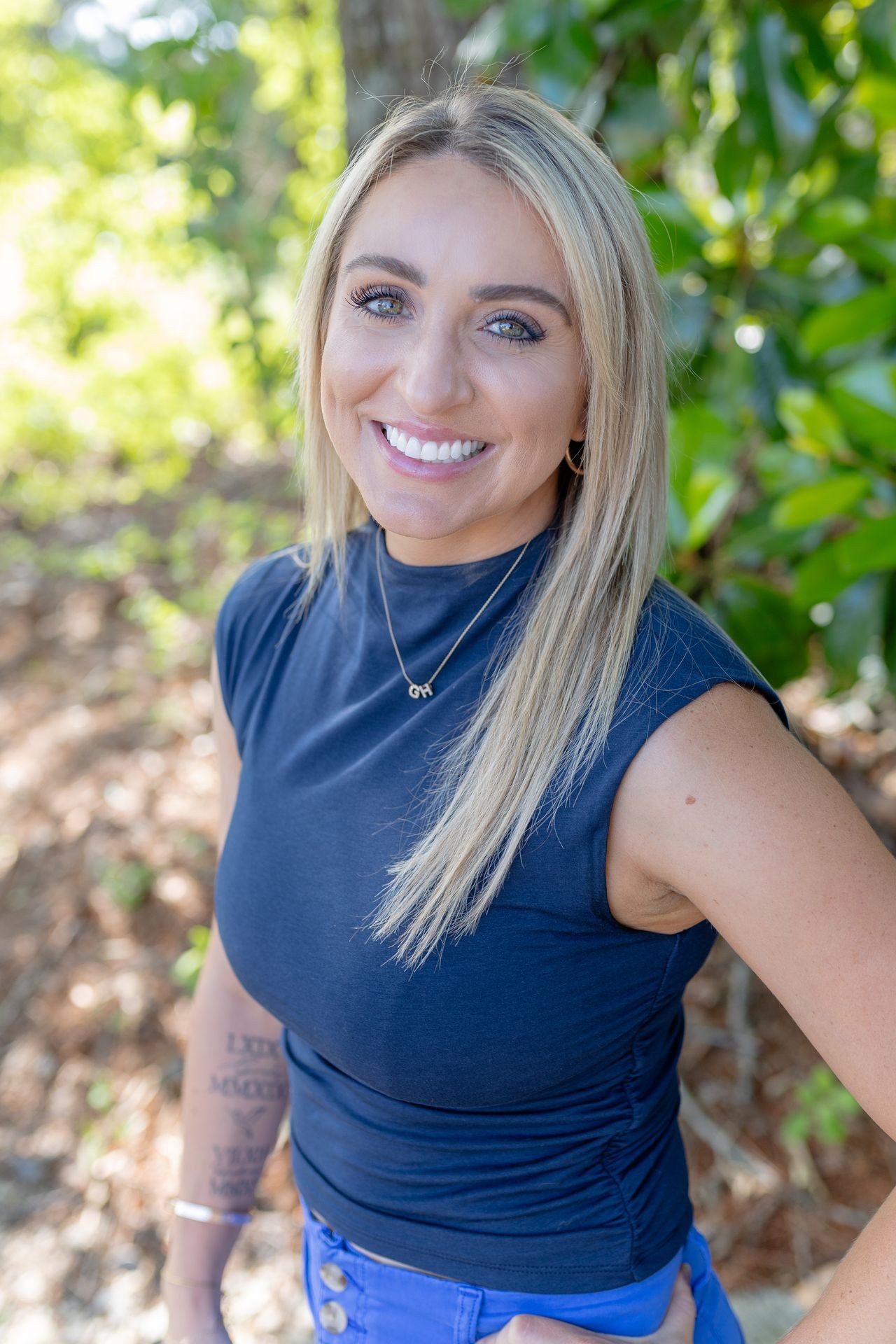 Smiling woman with long blonde hair in a sleeveless navy top stands outdoors near greenery.