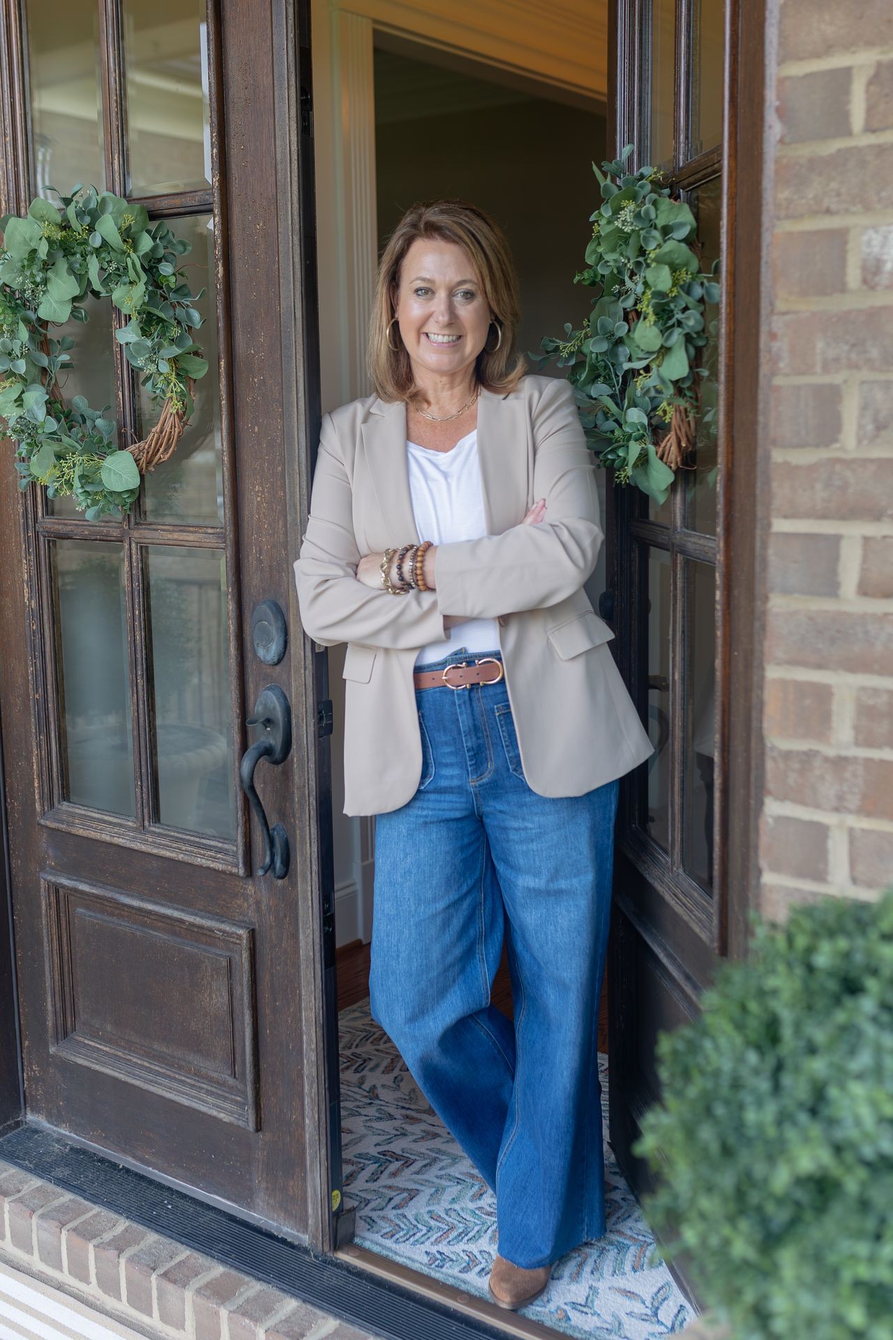 Smiling woman in casual attire leaning against a door with wreaths, standing on patterned mat.