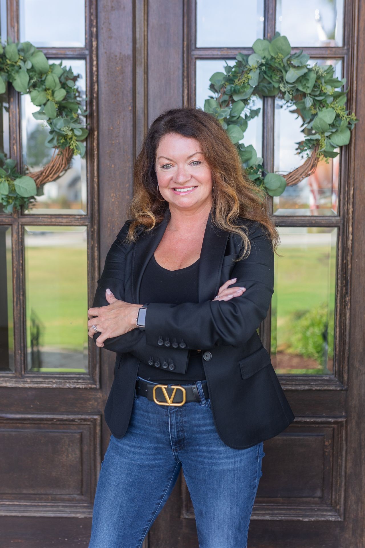 Woman in black blazer and jeans standing with arms crossed in front of a door with wreaths.