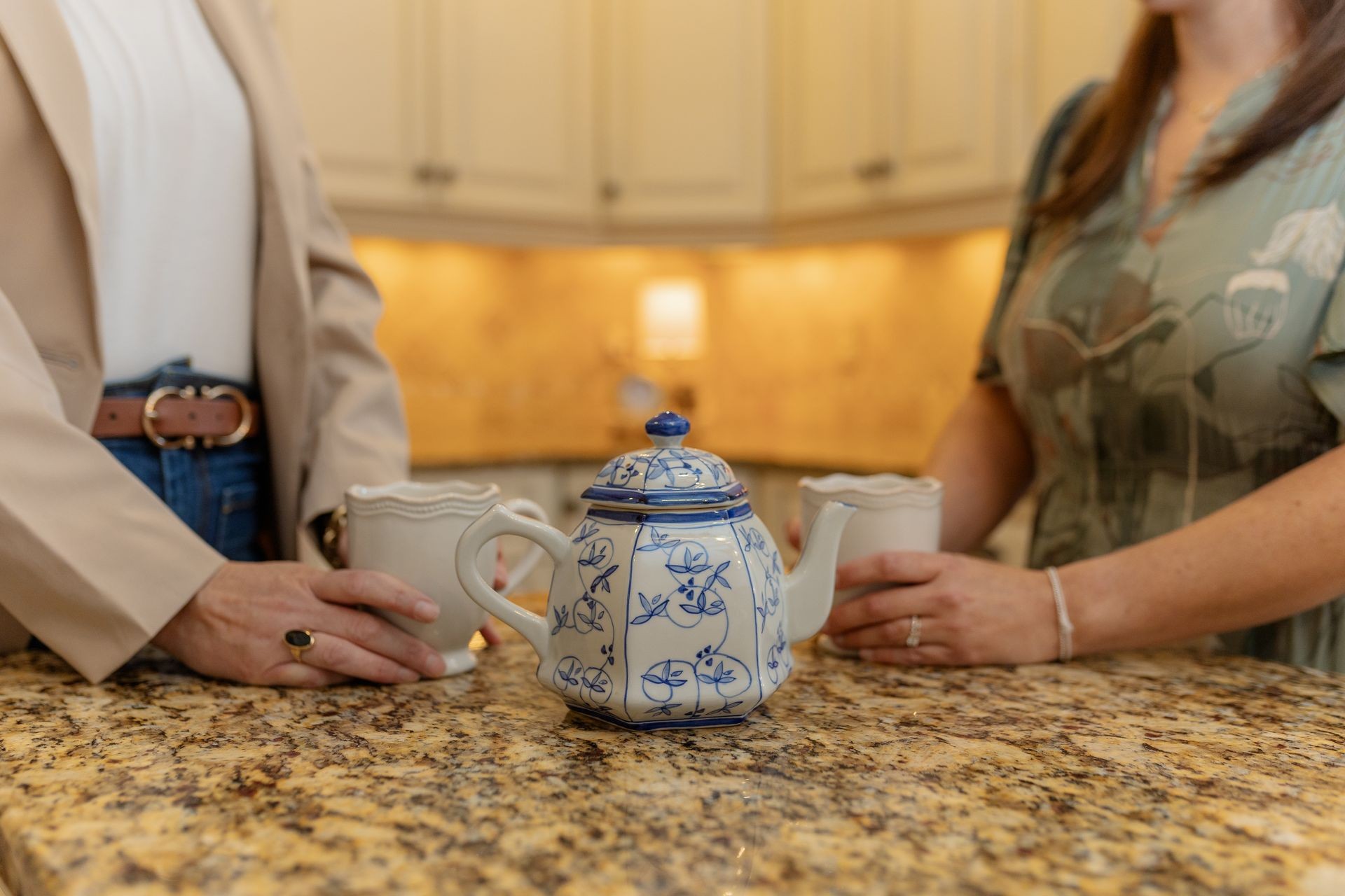 Two people holding mugs near a blue and white teapot on a granite countertop.