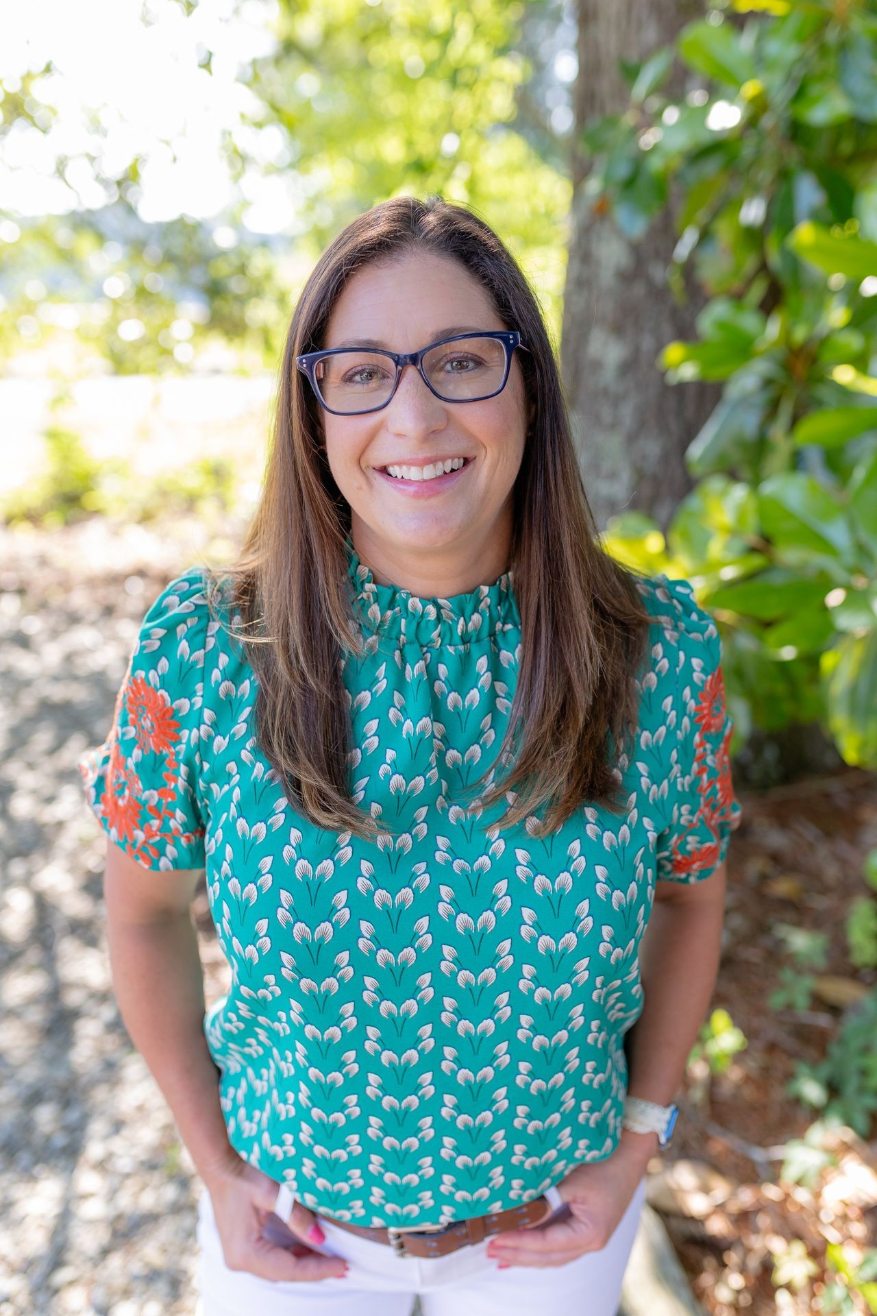 Person wearing a green and orange patterned blouse and white pants standing outdoors with greenery in the background.