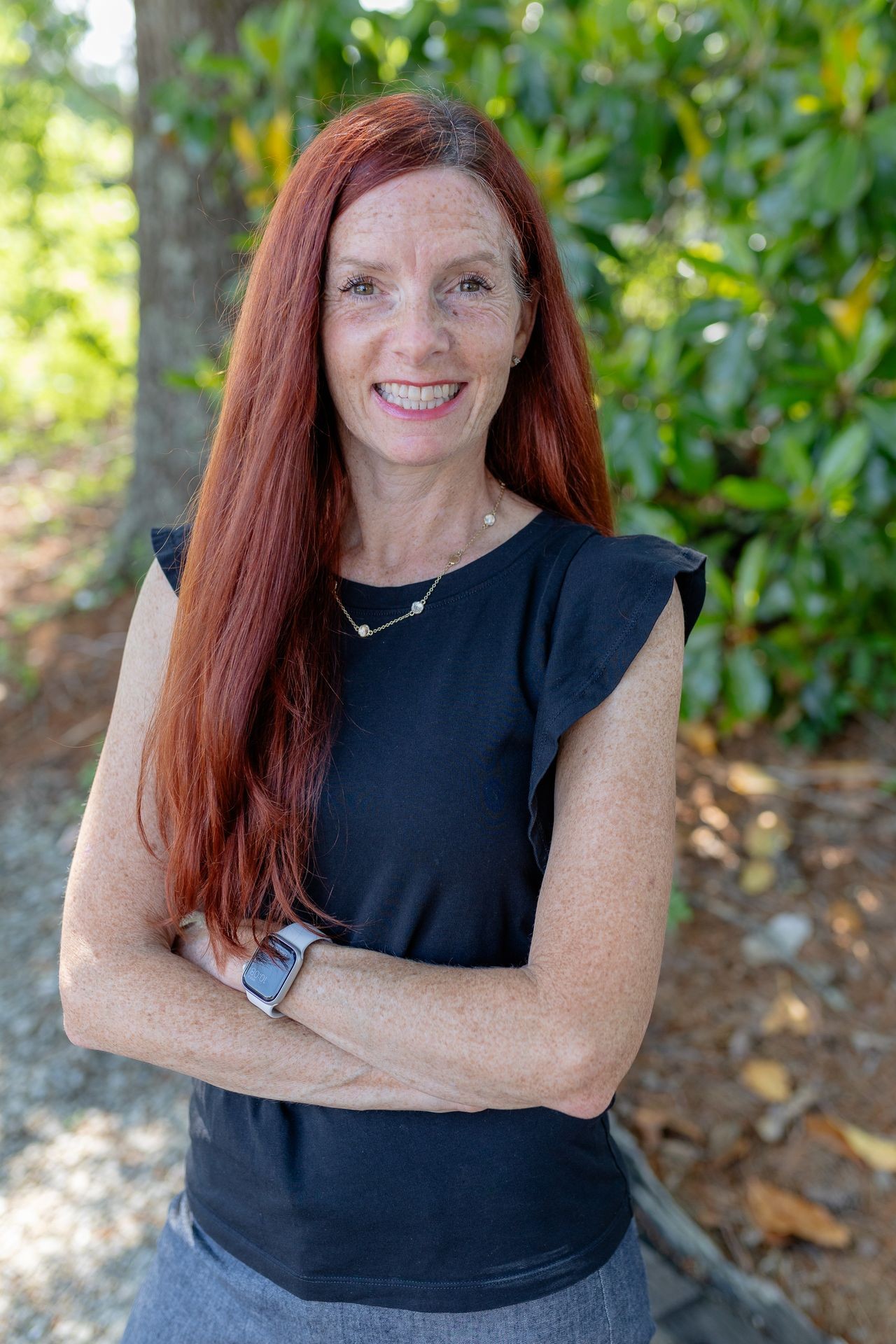 Woman with long red hair smiling, standing outdoors with green foliage background.