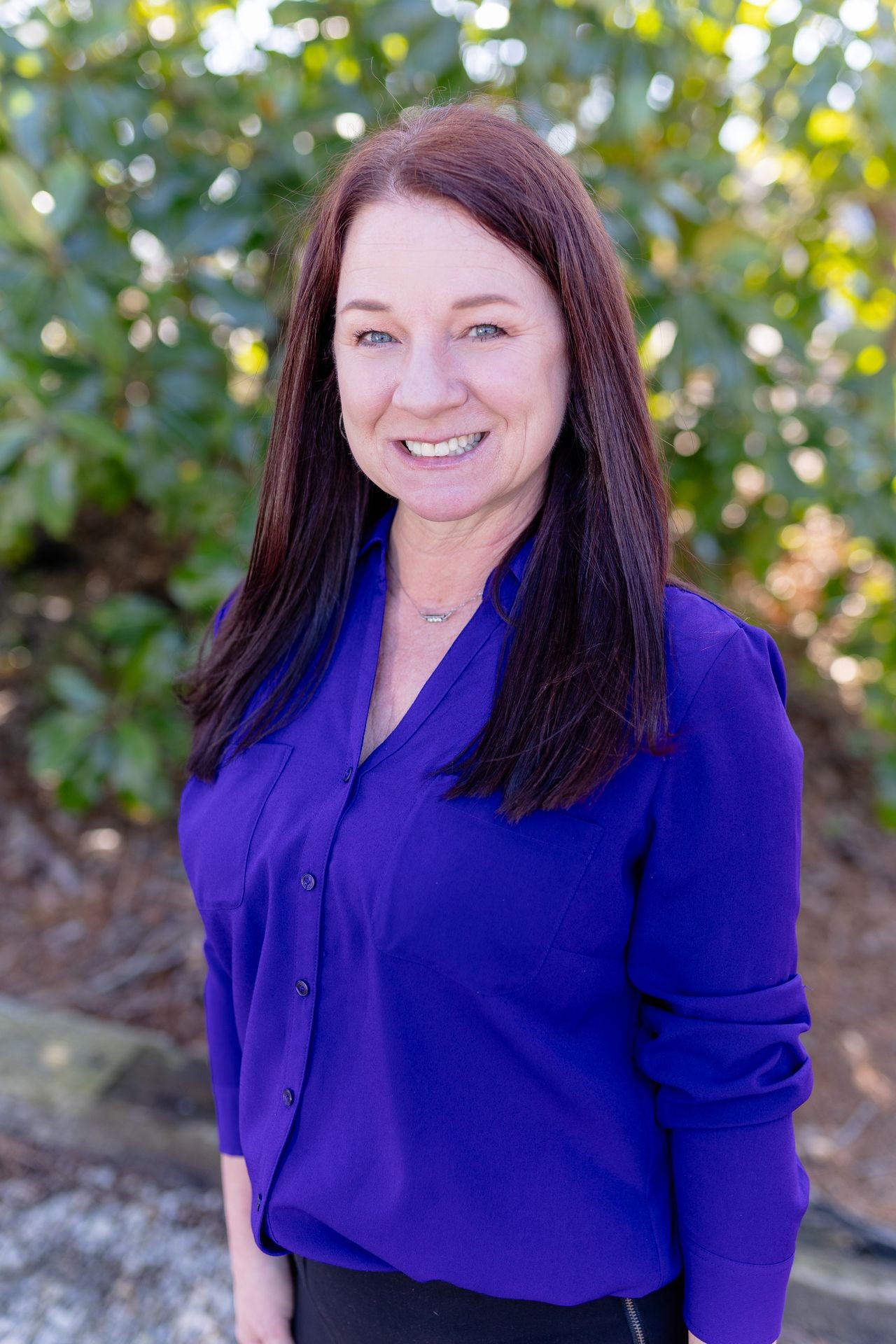 Smiling woman with long hair in a purple blouse, standing outdoors with greenery in the background.