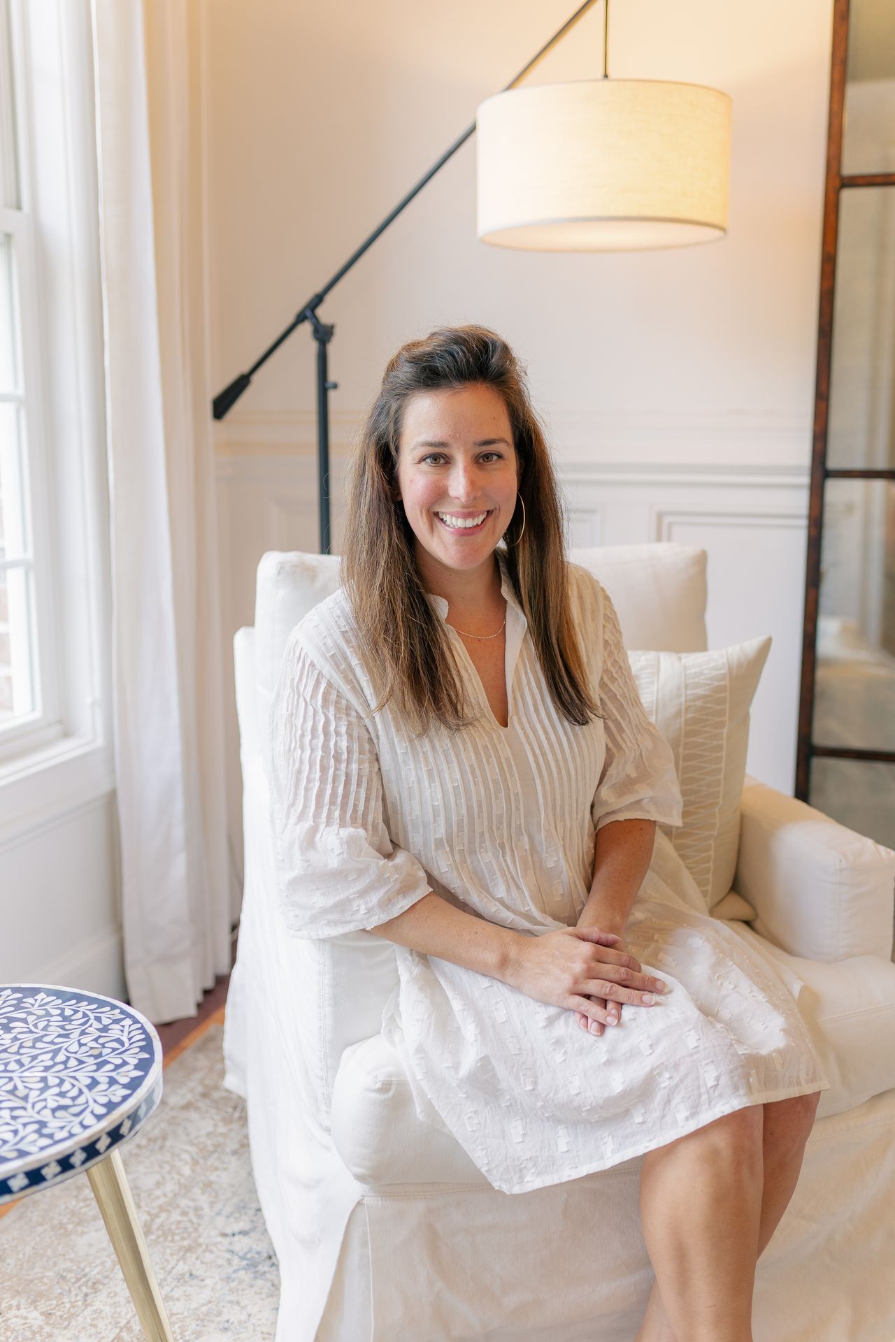 Smiling woman in a white dress seated in a bright room with a floor lamp and window.