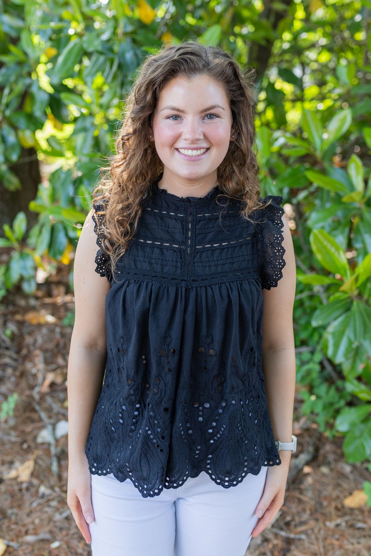 Smiling person in a black sleeveless top standing outdoors in front of green foliage.