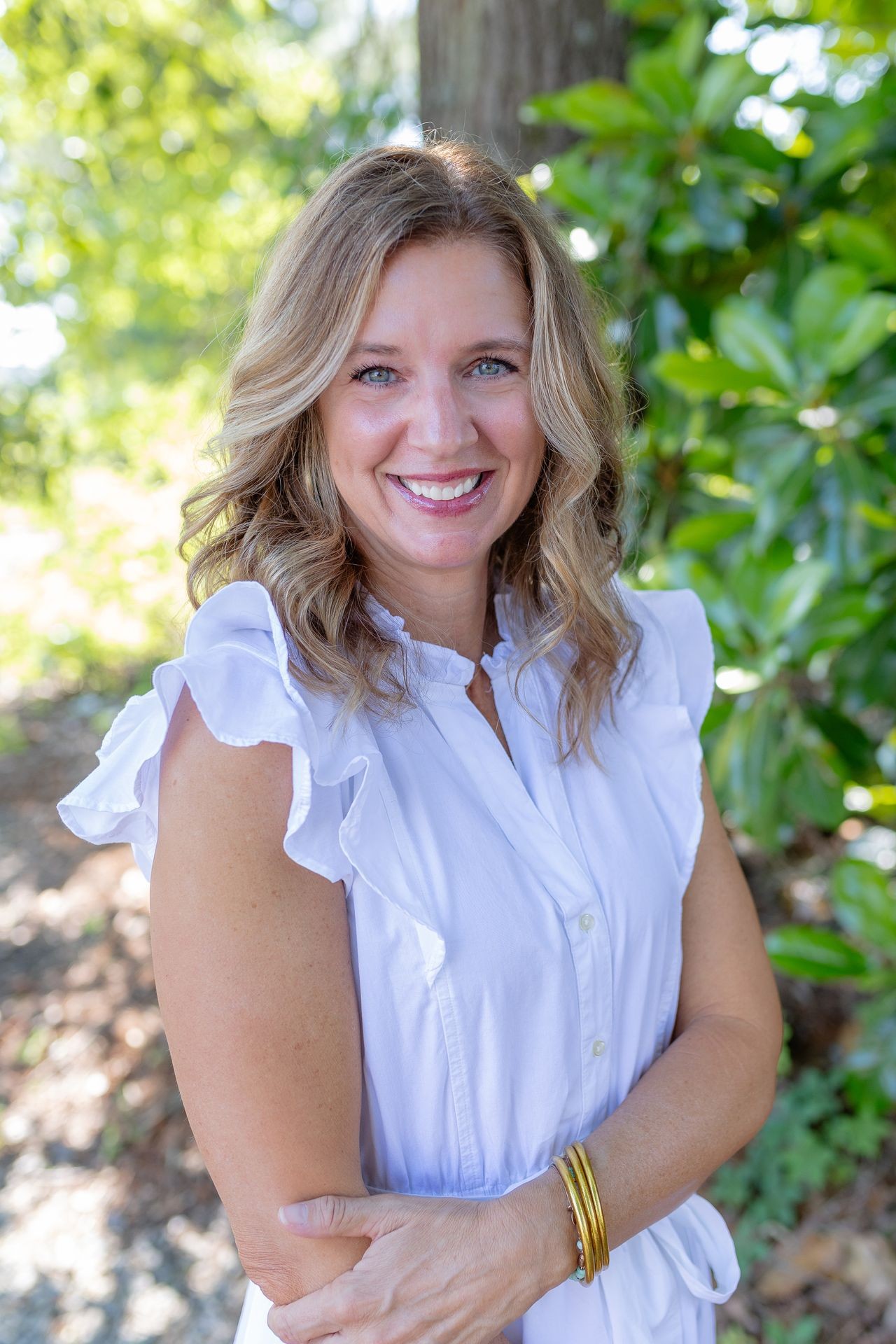 Smiling woman in a white dress standing outdoors with greenery in the background.