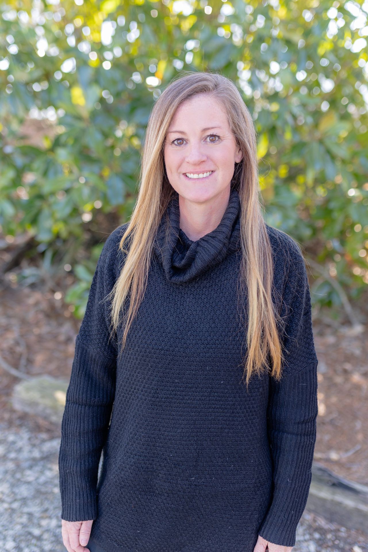 Person wearing a black sweater standing in front of green foliage.