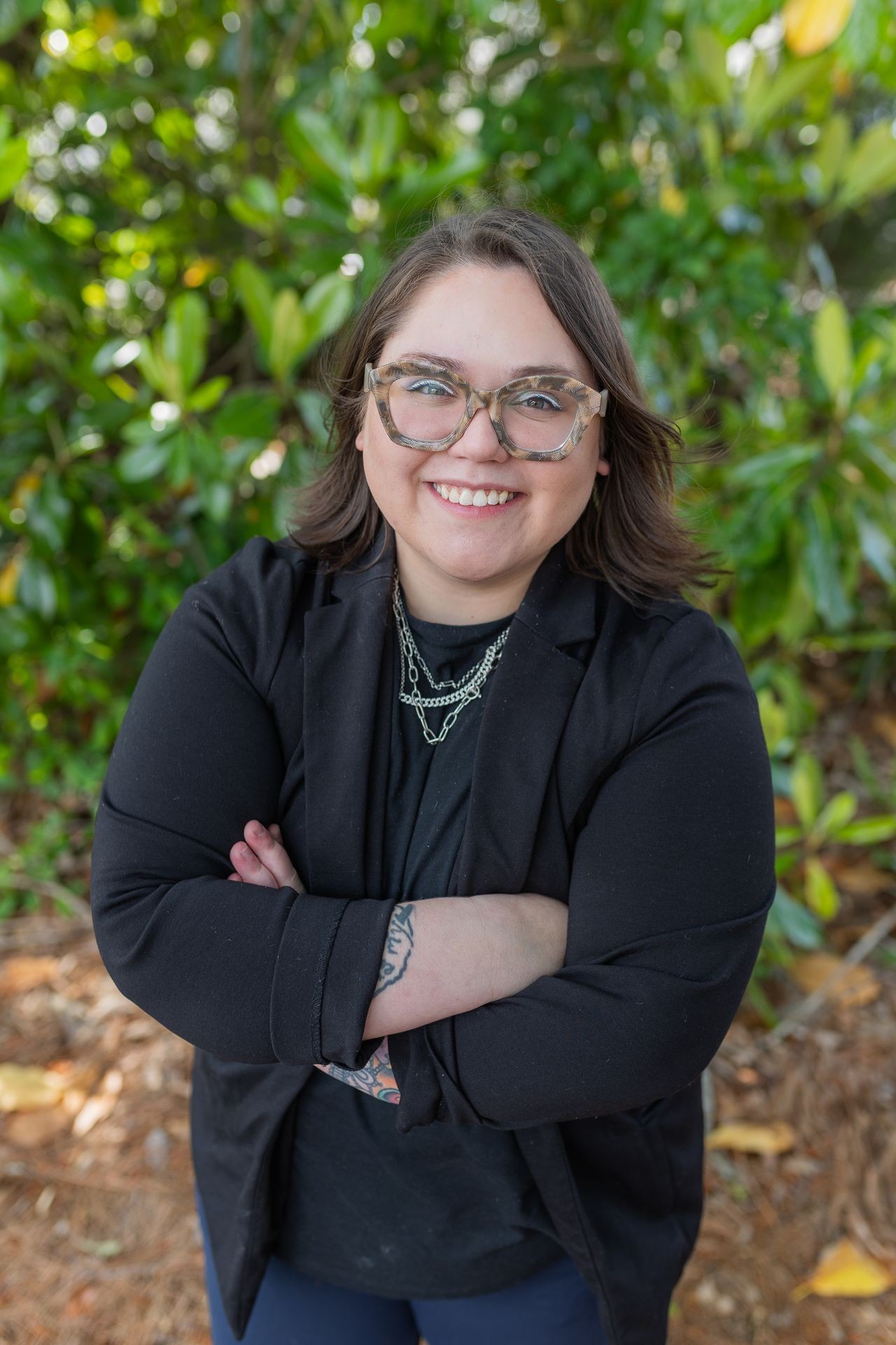 Person smiling with glasses and folded arms, standing outdoors with green foliage in the background.