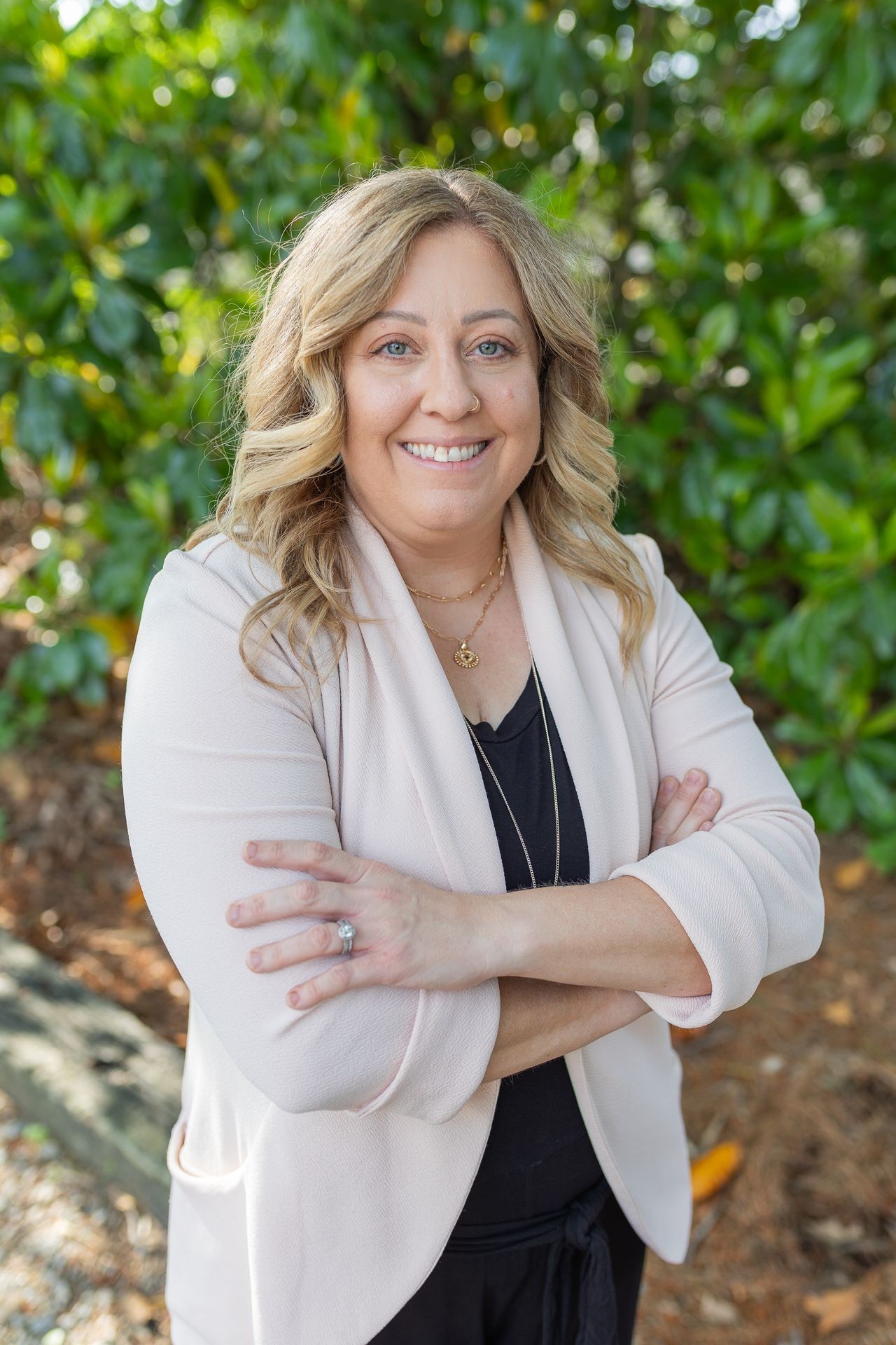 Smiling person with curly hair and blazer standing outside with arms crossed.