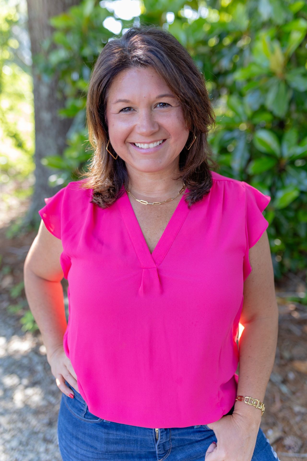 Person wearing a bright pink blouse and blue jeans, standing outside with green foliage in the background.