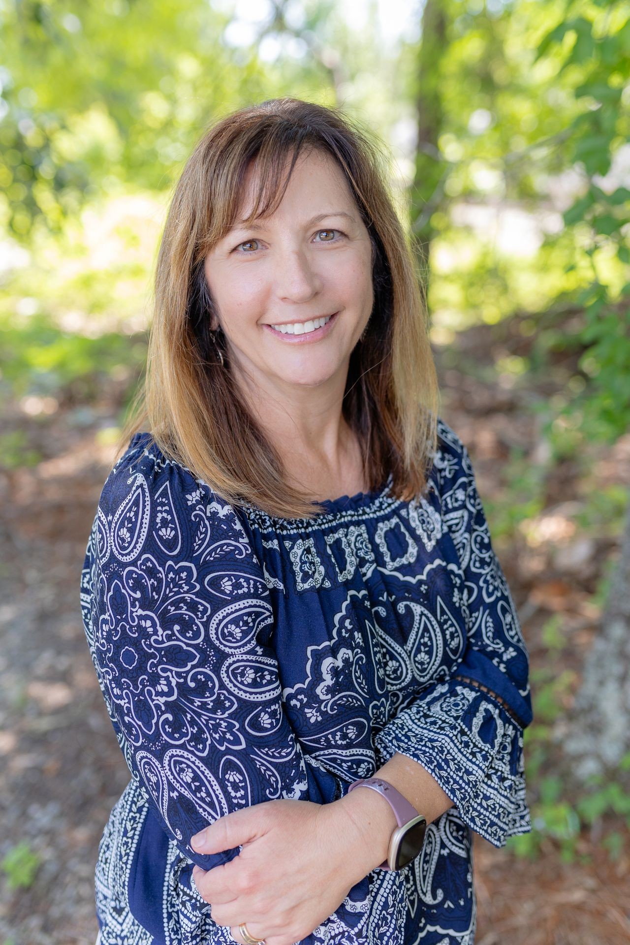 Person wearing a blue patterned blouse with arms crossed, standing outdoors surrounded by green foliage.