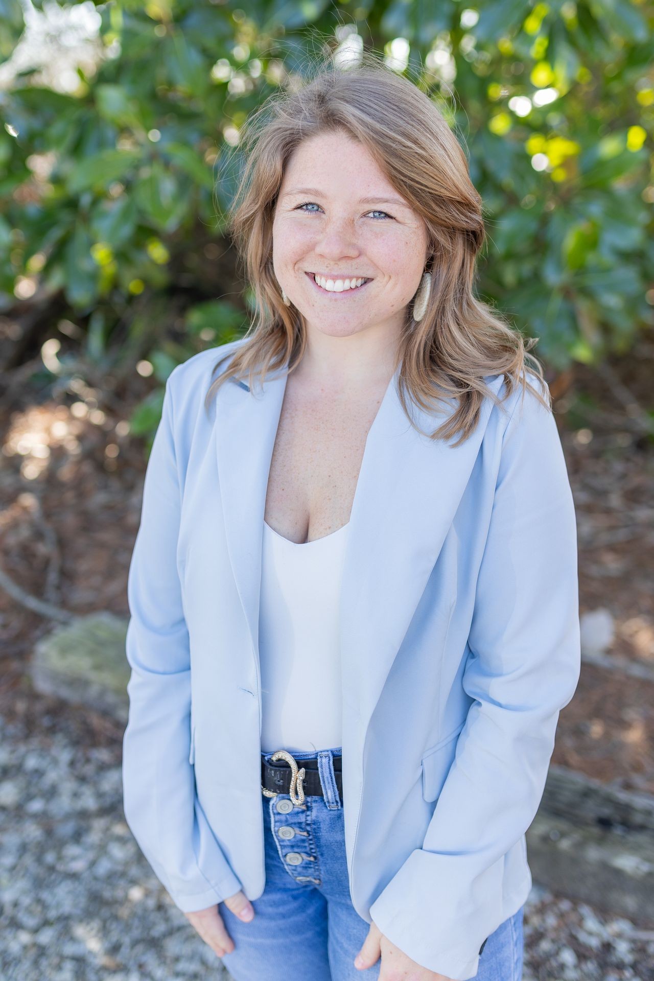 Person wearing a light blue blazer and blue jeans stands outside with blurred green foliage in the background.