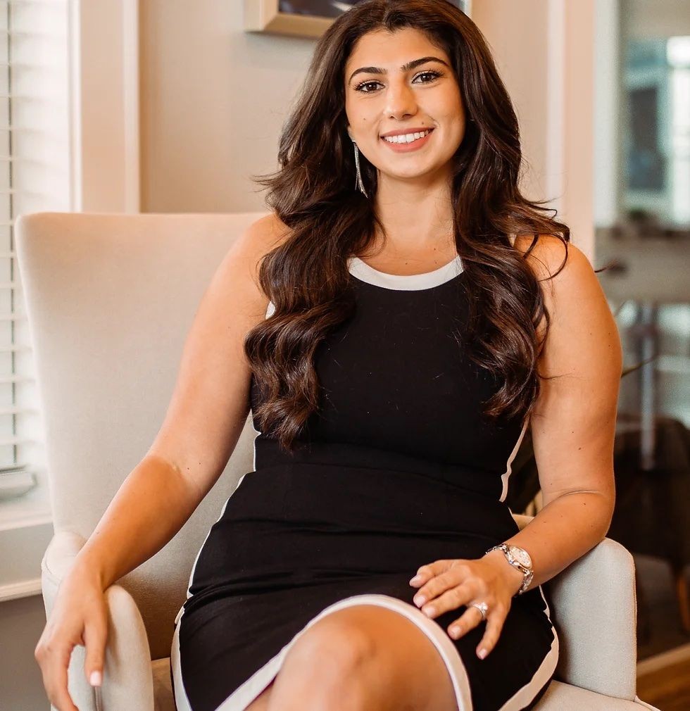 Woman with long hair sitting on a chair, wearing a black dress with white trim and smiling.