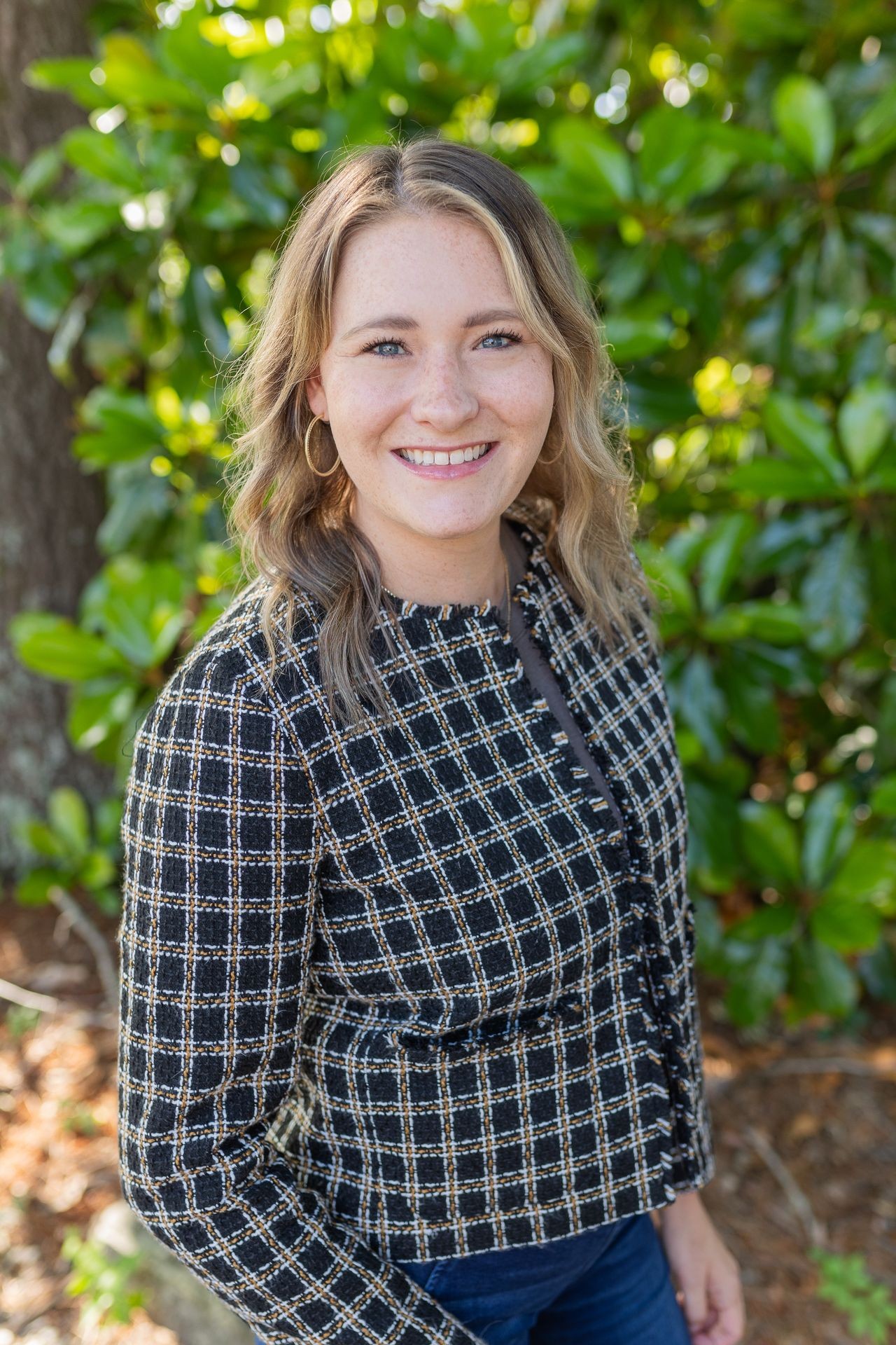 Woman smiling outdoors with a patterned jacket, standing in front of green foliage.