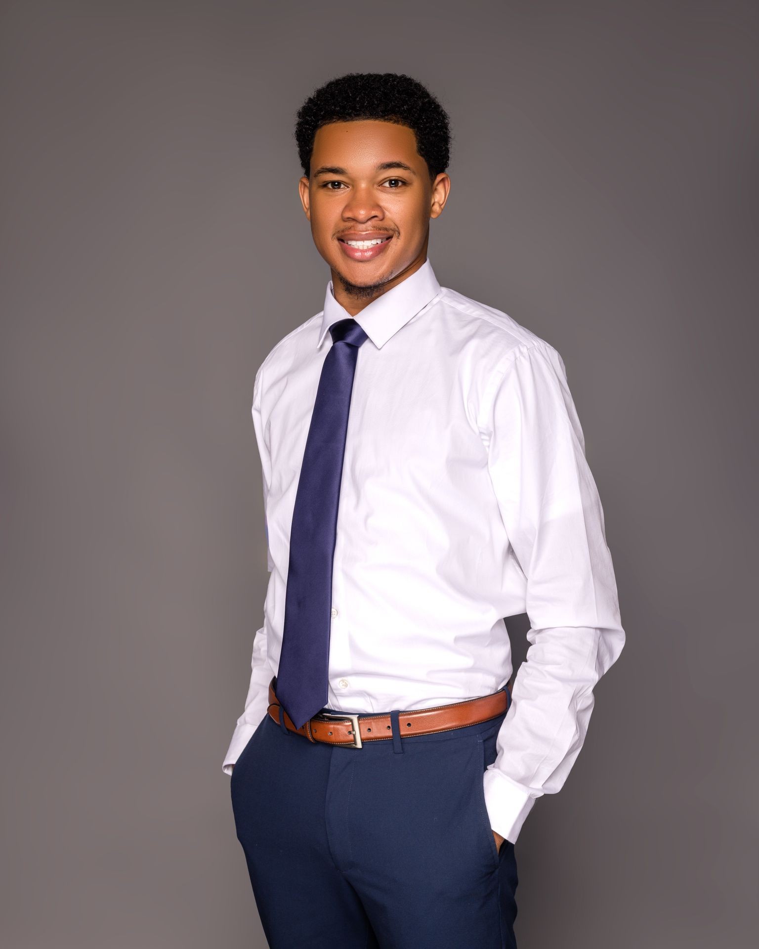 Man in white shirt and blue tie smiling with hands in pockets against grey background.