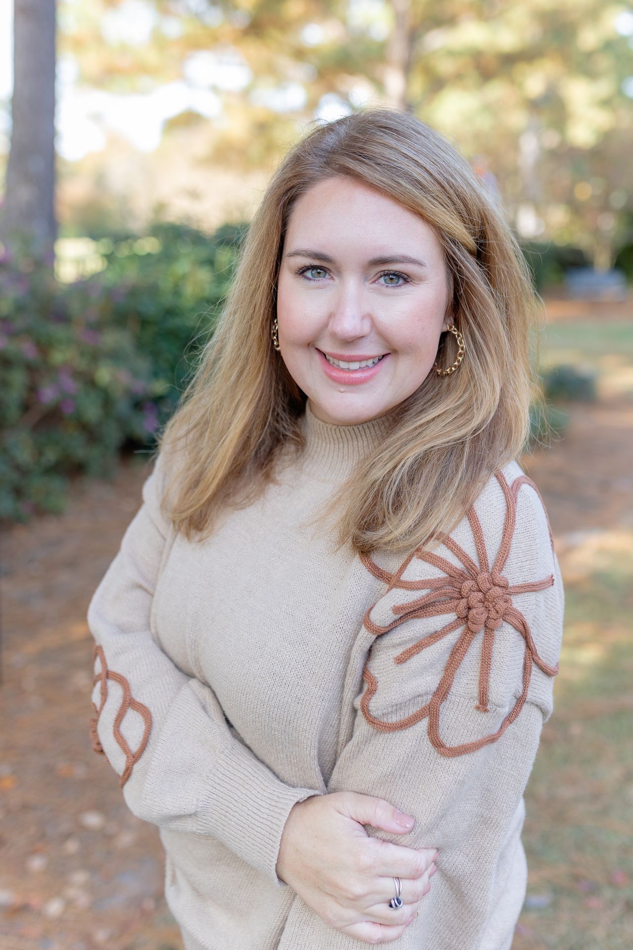 Person wearing a beige sweater with brown floral embroidery standing outdoors in a garden.