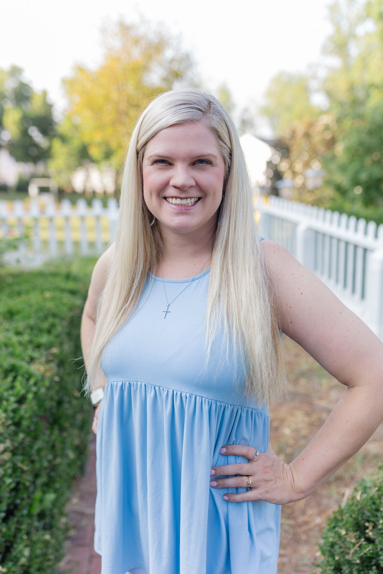 Woman in a blue dress smiling outdoors by a white fence and greenery.