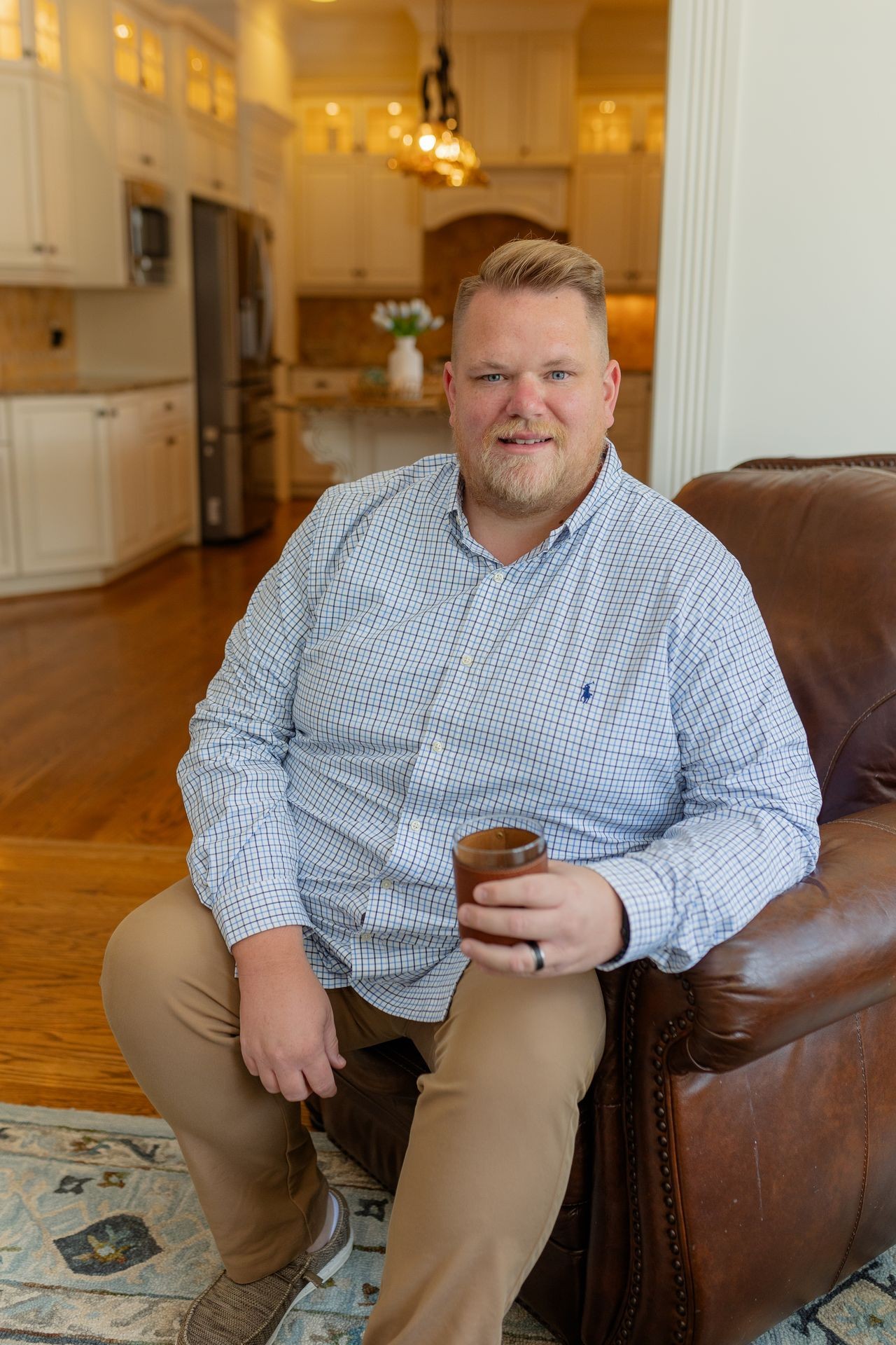 Individual sitting on a brown leather couch holding a cup, with a modern kitchen in the background.