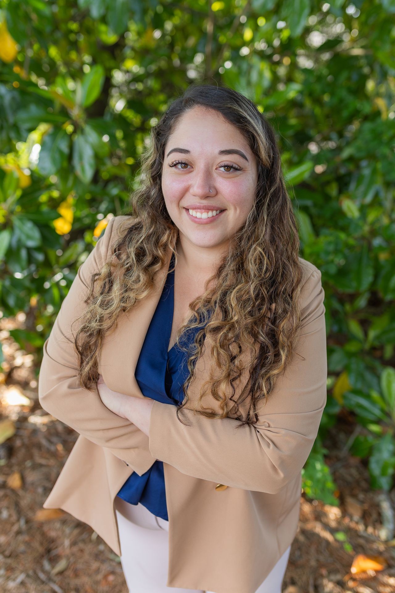 Smiling woman with curly hair in a beige blazer standing outdoors against a background of green foliage.