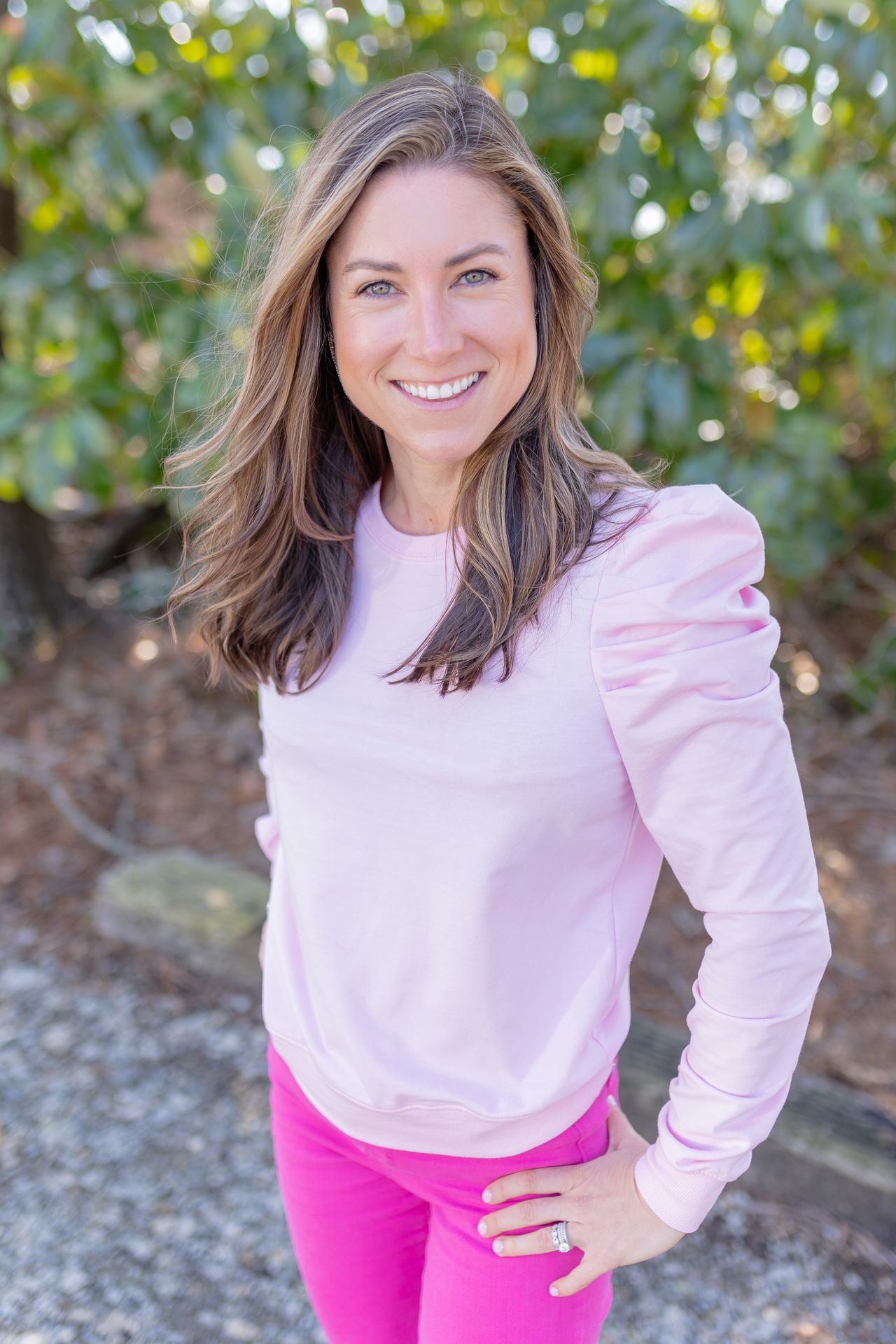 Woman in pink outfit smiles outdoors with a blurred green foliage background.